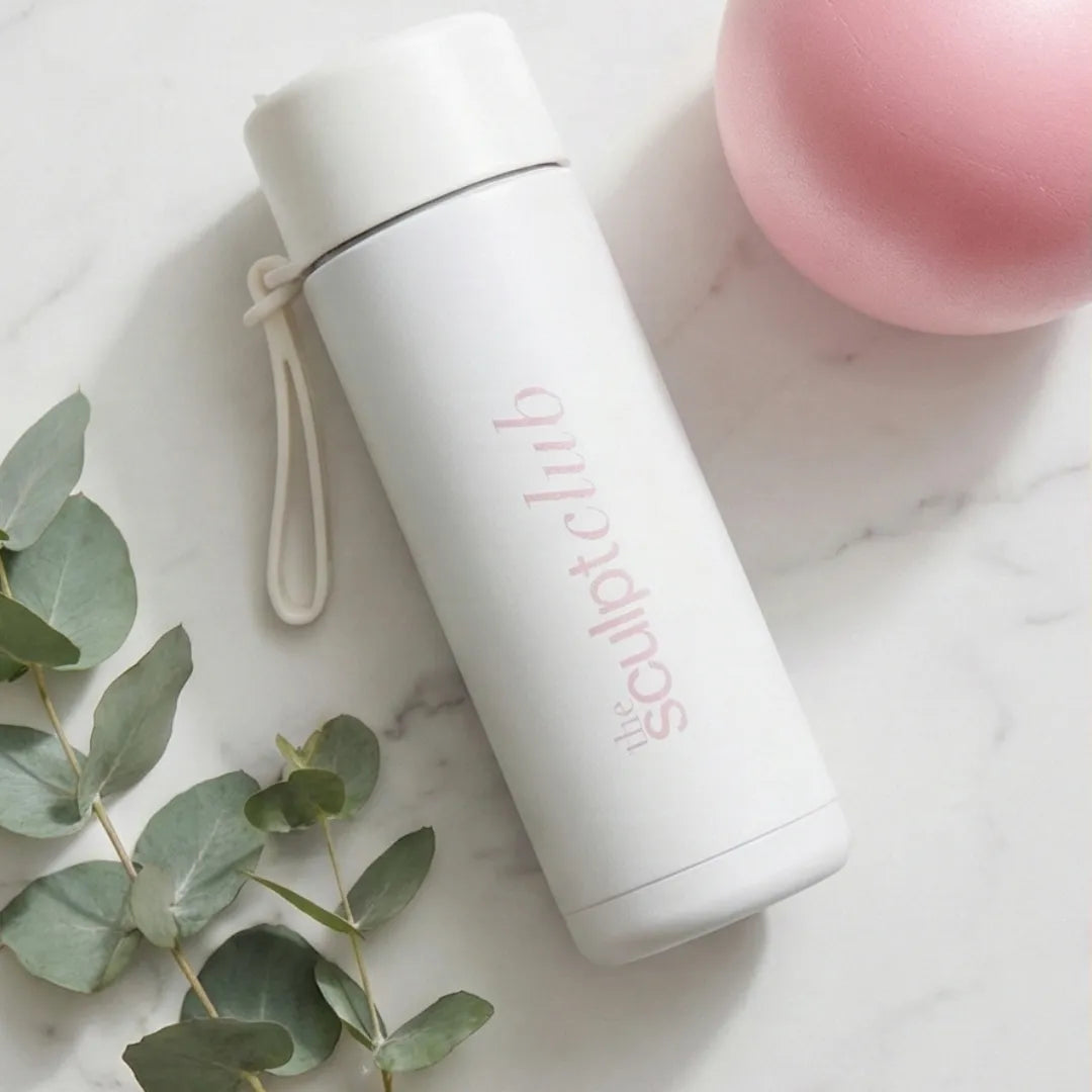 White insulated bottle with 'the sculpt club' branding on a marble surface with eucalyptus leaves and a pink pilates ball.