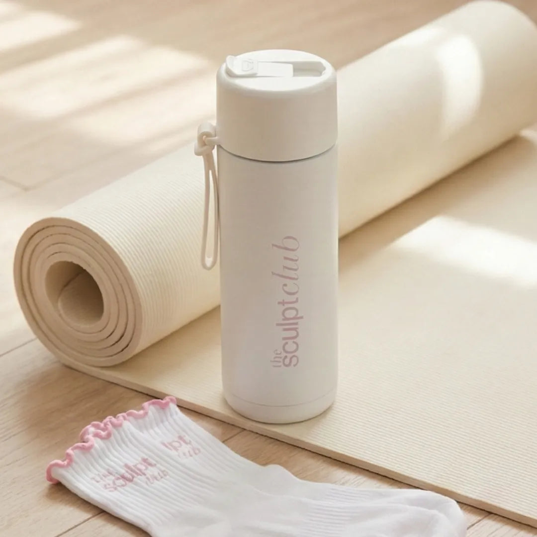 White water bottle with 'sculpt club' branding on a wooden floor with a yoga mat and socks.